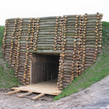 Aus Eichenstämmen ist eine steile Wand errichtet. In der Mitte befindet sich ein tunnelartiger Durchgang, dessen Boden mit Holzbohlen belegt ist. Rechts und links geht die steile Wand in eine grasbewachsene Böschung über. (© LAKD M-V/LA)